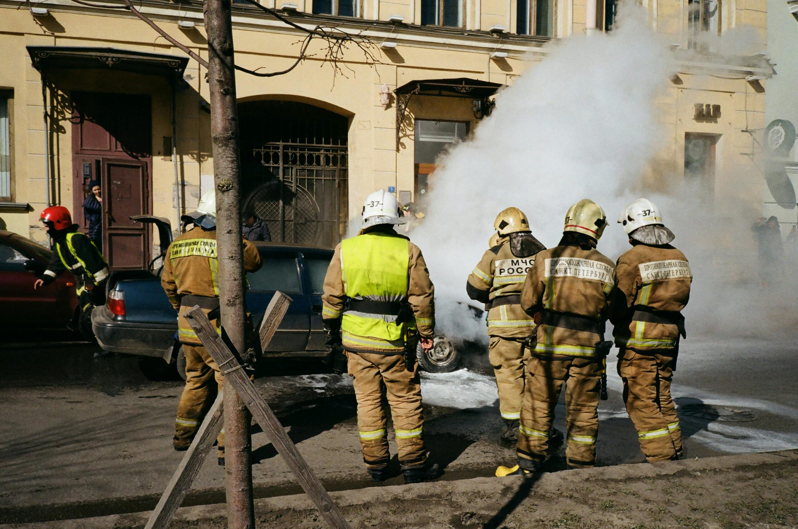 Firefighters responding to a car fire on a street in St. Petersburg, Russia, with smoke and safety gear in view.